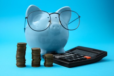 Photo of Piggy bank in glasses, stacks of coins and calculator on light blue background