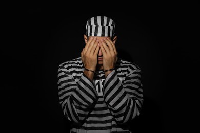Photo of Remorseful prisoner in striped uniform with handcuffs hiding his face on black background