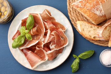 Photo of Slices of tasty cured ham, basil, olives and bread on blue wooden table, flat lay