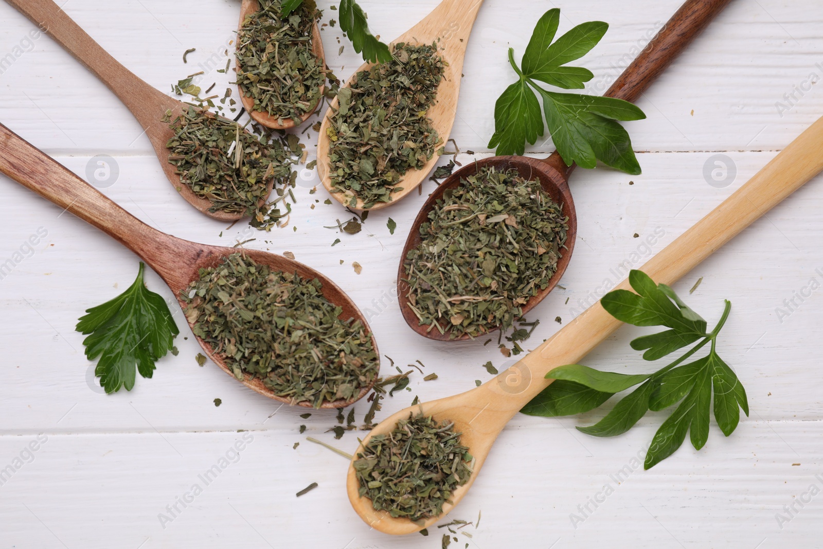 Photo of Spoons with dried aromatic parsley and fresh leaves on white wooden table, flat lay