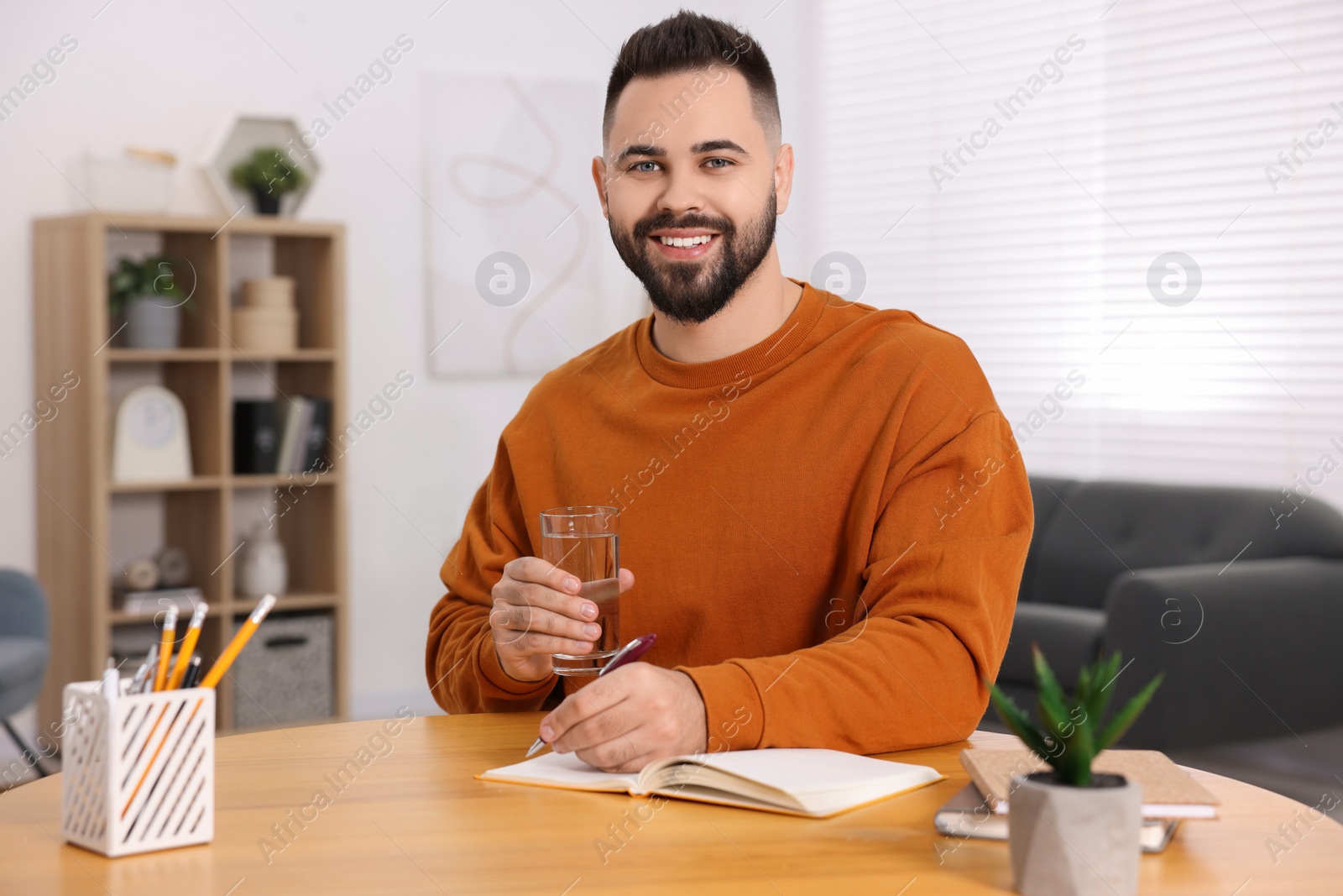 Photo of Young man with glass of water writing in notebook at wooden table indoors