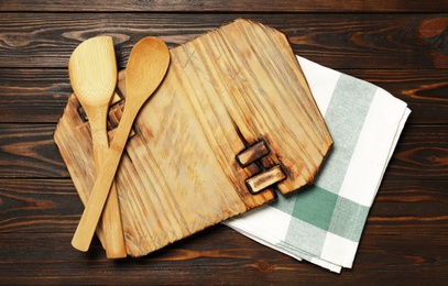 Photo of Board, spatula and spoon with towel on brown wooden table, flat lay. Cooking utensils
