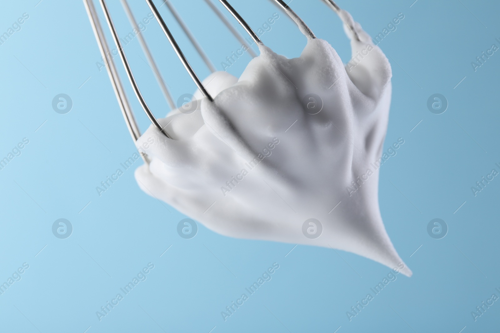 Photo of Whisk with whipped cream on light blue background, closeup