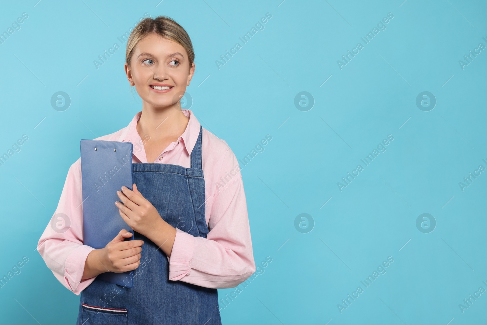 Photo of Beautiful young woman in denim apron with clipboard on light blue background. Space for text