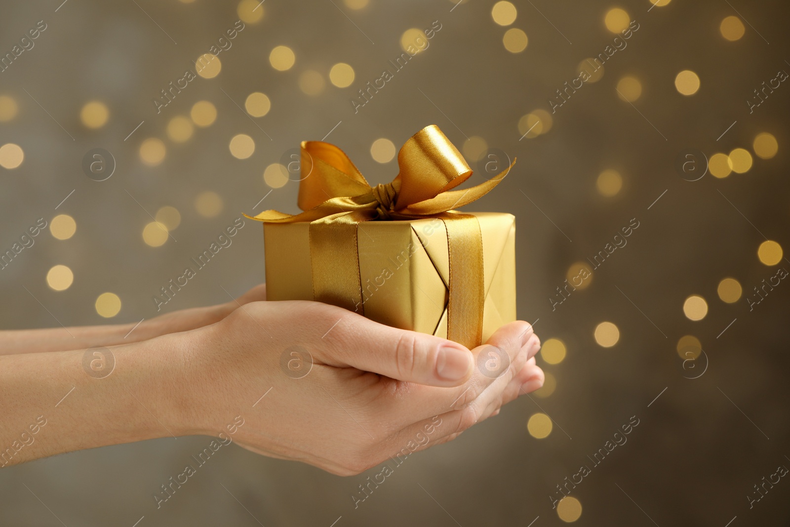 Photo of Woman holding beautifully wrapped gift box against blurred festive lights, closeup