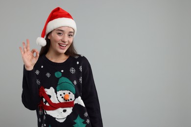 Photo of Happy young woman in Christmas sweater and Santa hat showing OK gesture on grey background. Space for text
