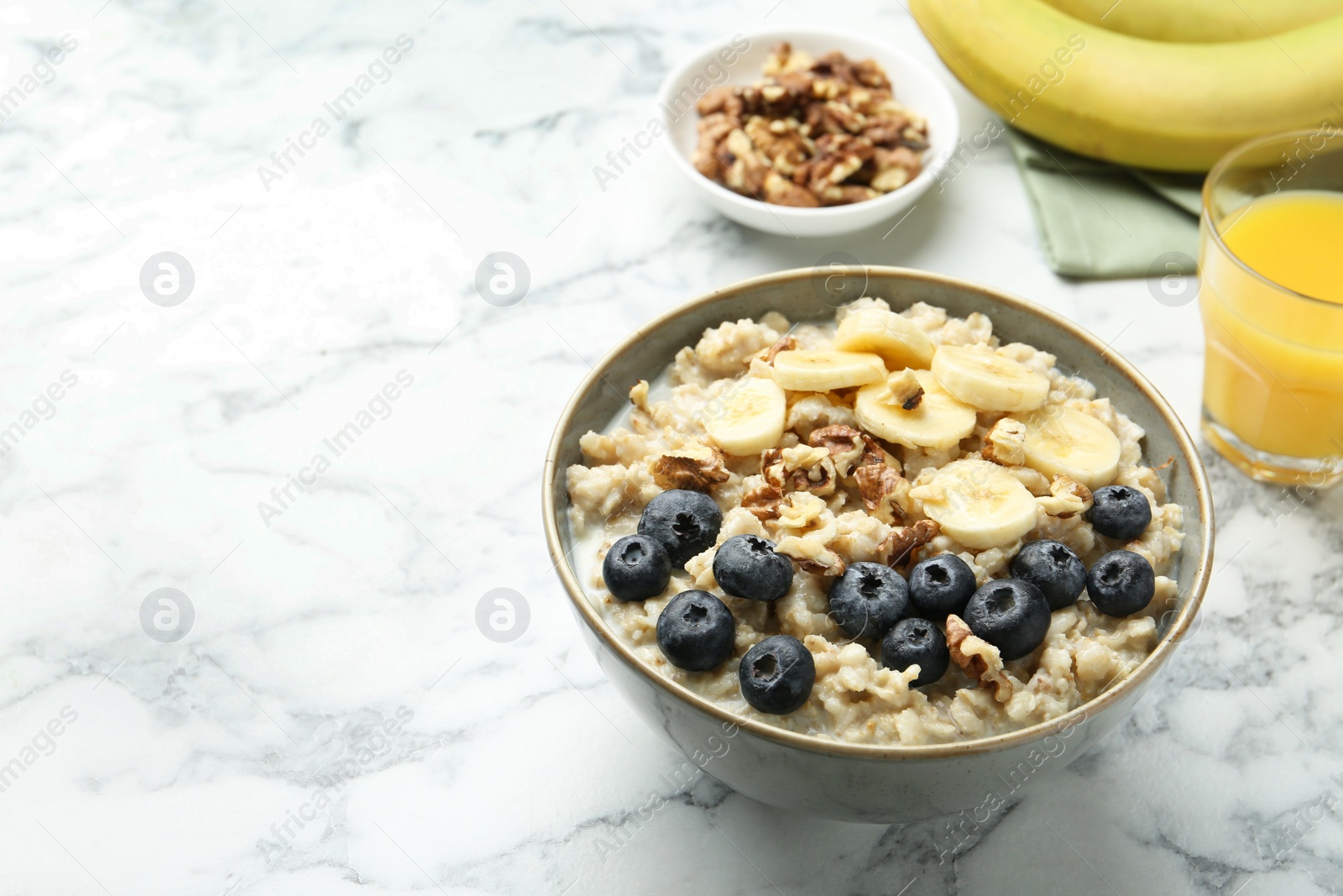 Photo of Tasty oatmeal with banana, blueberries, walnuts and milk served in bowl on white marble table, space for text