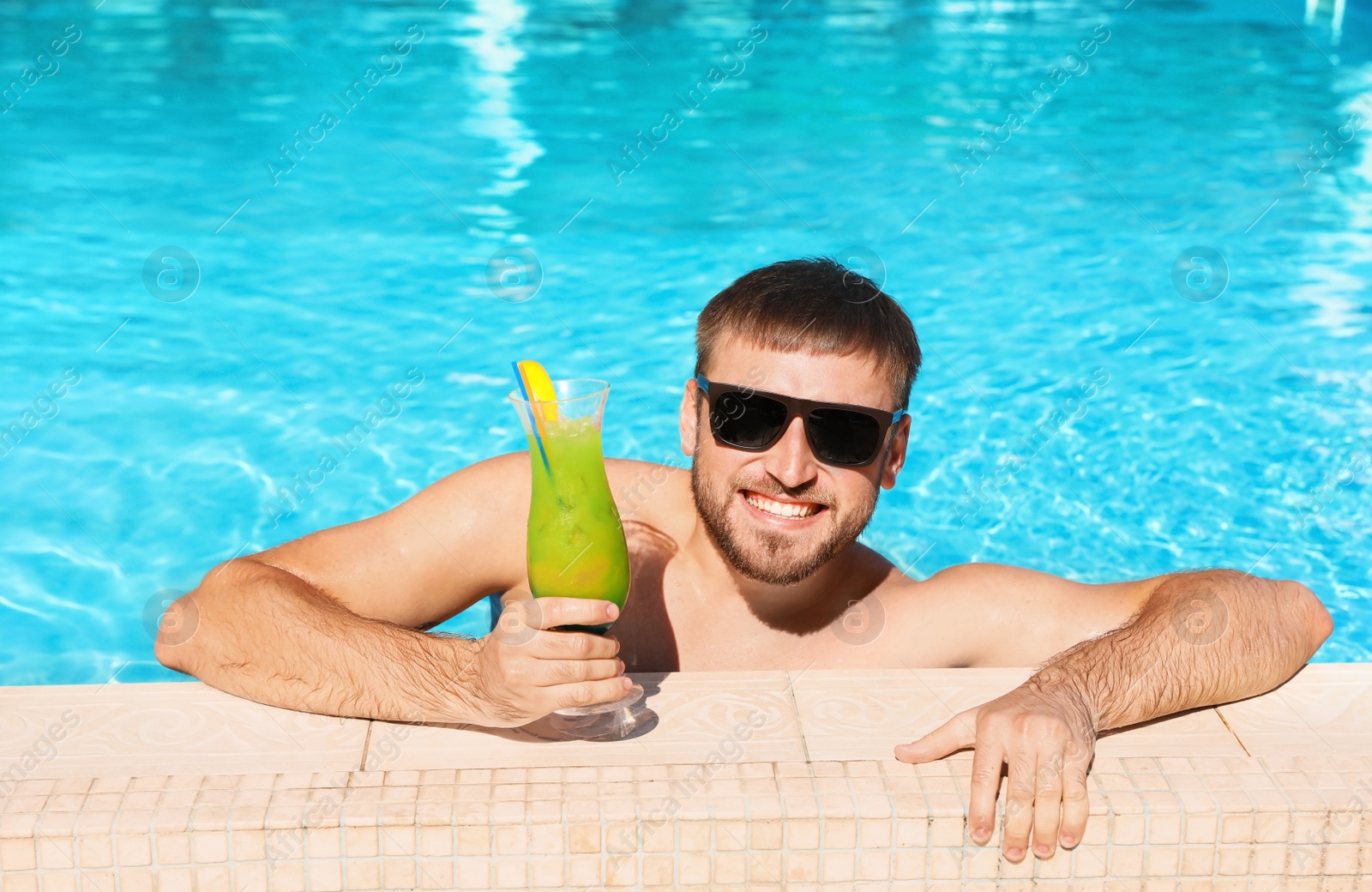 Photo of Young bearded man with refreshing cocktail in swimming pool at resort