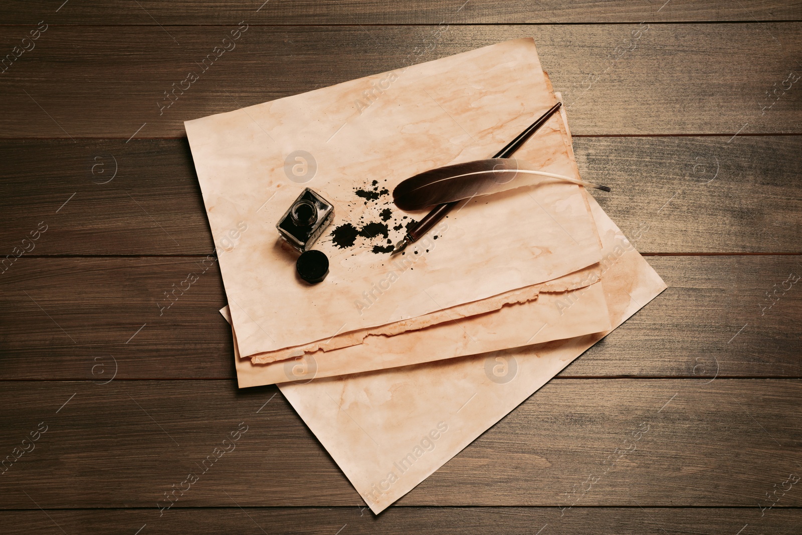 Photo of Feather, fountain pen, inkwell and vintage parchment with ink stains on wooden table, top view