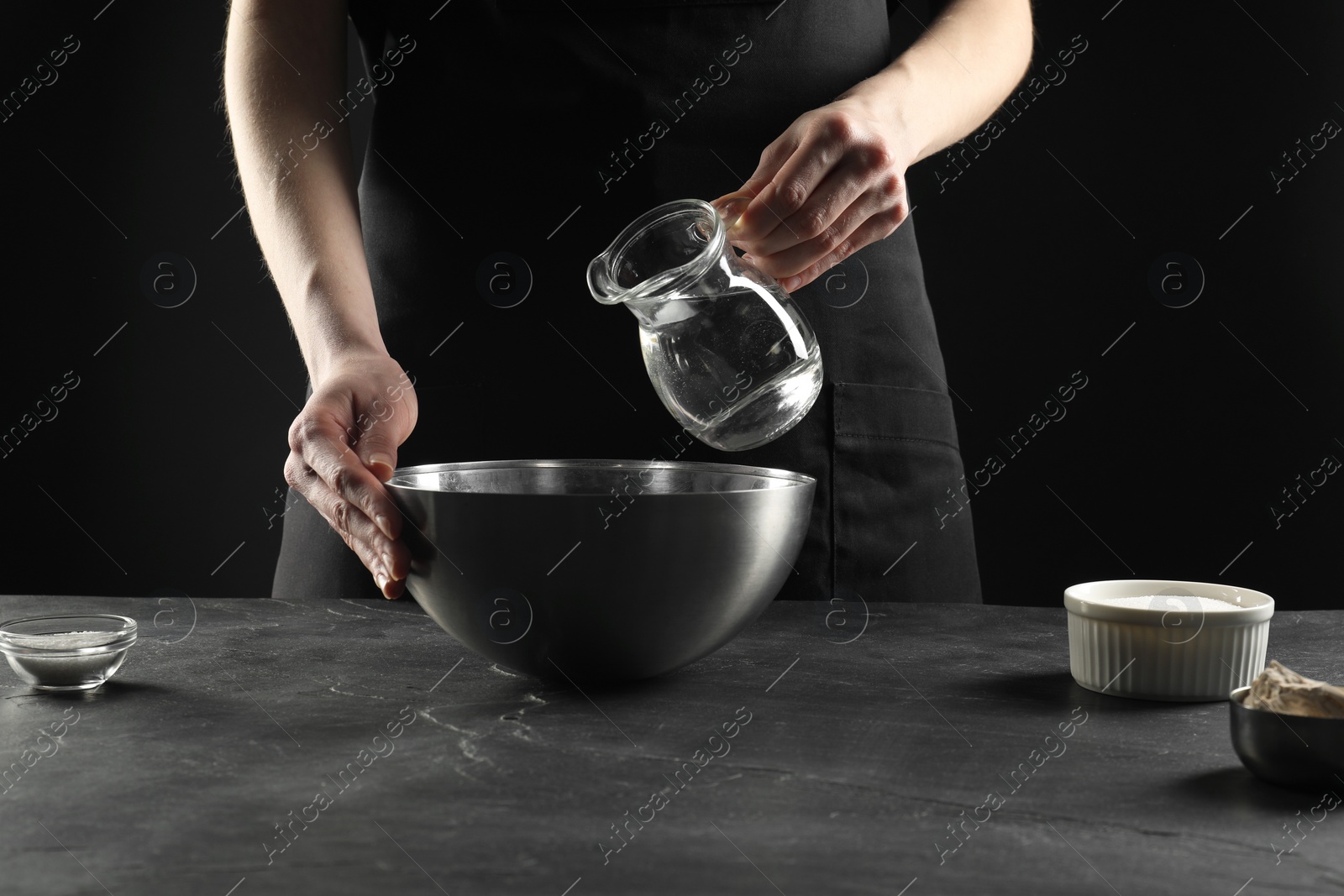 Photo of Making bread. Woman pouring water into bowl at grey textured table, closeup