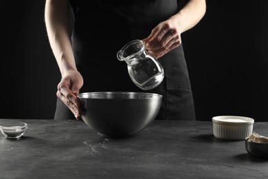 Photo of Making bread. Woman pouring water into bowl at grey textured table, closeup