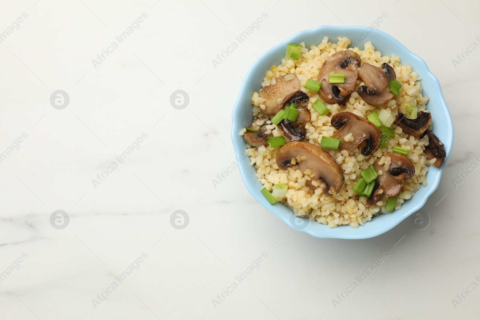 Photo of Delicious bulgur with mushrooms and green onion in bowl on white marble table, top view. Space for text