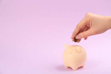 Woman putting coin into piggy bank on violet background, closeup. Space for text