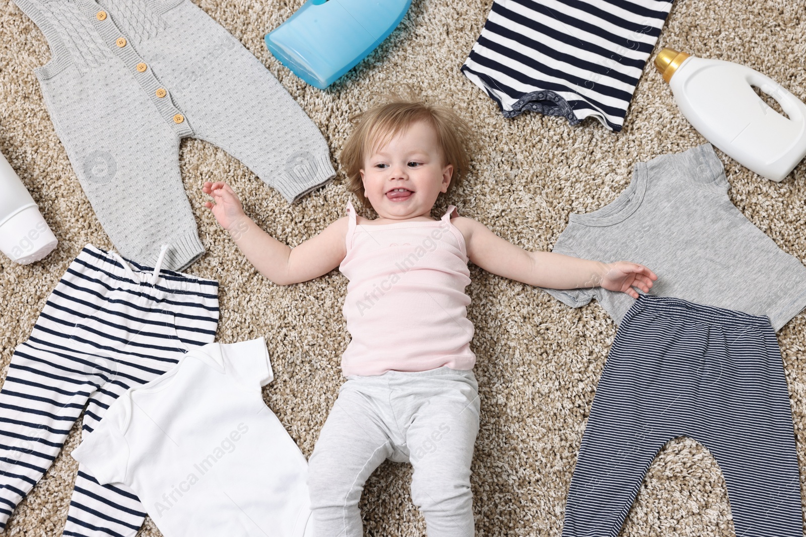 Photo of Little girl among baby clothes and detergents on carpet, top view