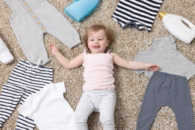 Little girl among baby clothes and detergents on carpet, top view