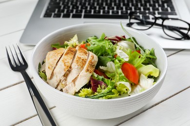 Photo of Bowl with tasty food, fork, laptop and glasses on white wooden table, closeup. Business lunch