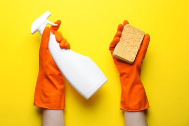 Photo of Woman in rubber gloves holding sponge and detergent on yellow background, top view