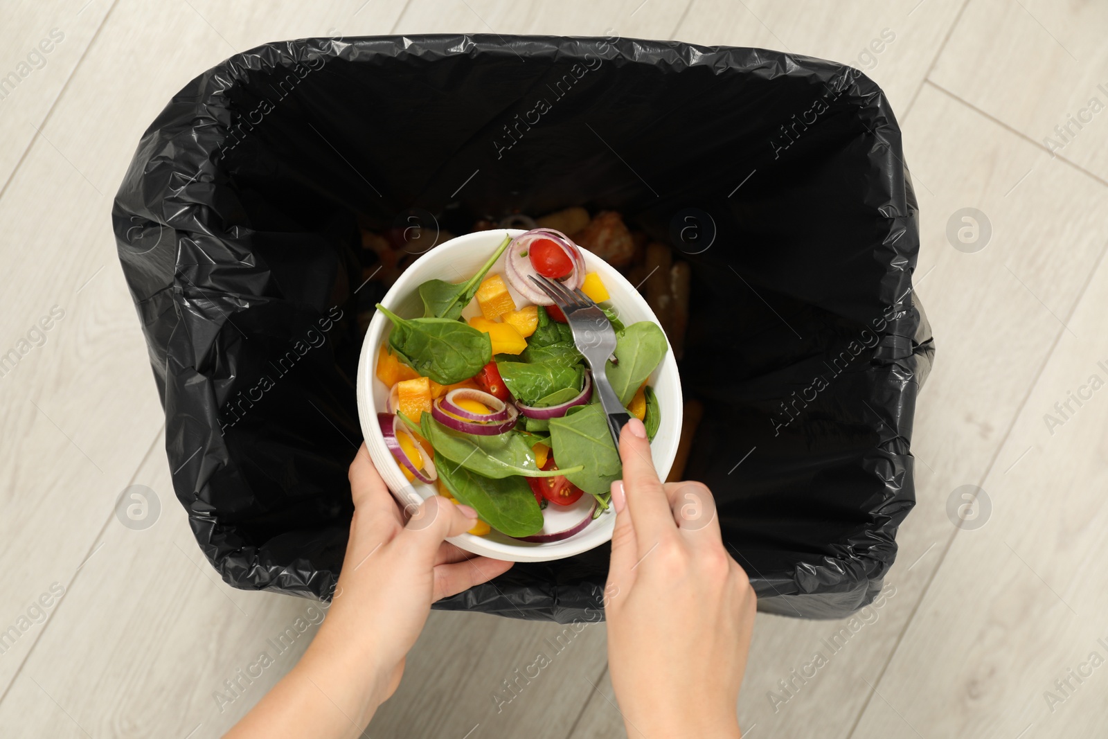 Photo of Woman throwing vegetable salad into bin indoors, top view