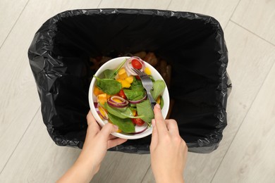 Woman throwing vegetable salad into bin indoors, top view