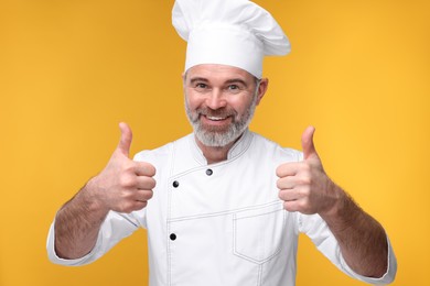 Photo of Happy chef in uniform showing thumbs up on orange background