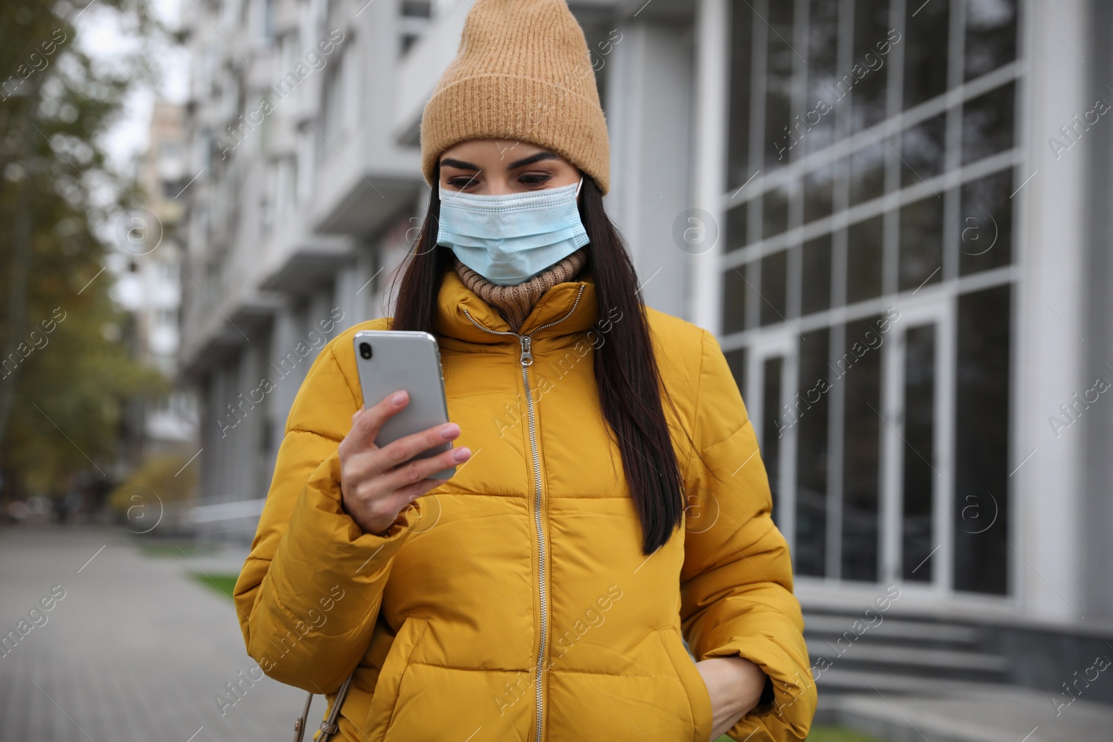 Photo of Young woman in medical face mask with smartphone walking outdoors. Personal protection during COVID-19 pandemic