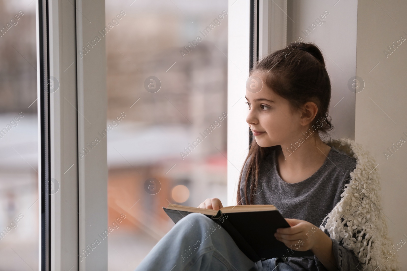 Photo of Cute little girl reading book near window at home. Space for text
