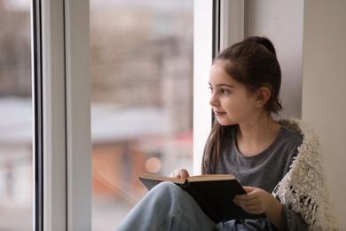 Cute little girl reading book near window at home. Space for text