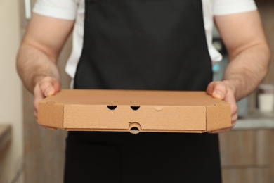 Photo of Waiter in apron with pizza box indoors, closeup. Food delivery service
