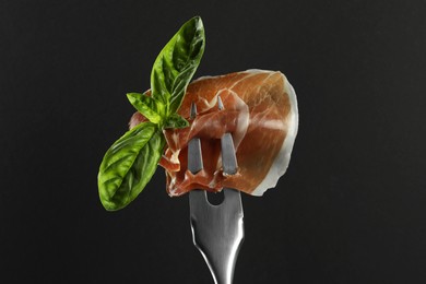 Photo of Fork with slice of tasty jamon and basil leaves on black background, closeup