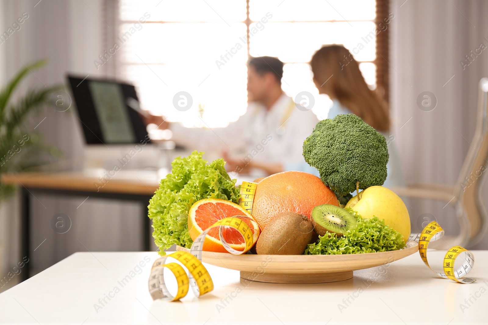 Photo of Nutritionist consulting patient at table in clinic, focus on plate with fruits, vegetables and measuring tape