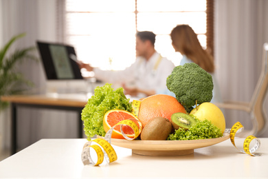 Nutritionist consulting patient at table in clinic, focus on plate with fruits, vegetables and measuring tape