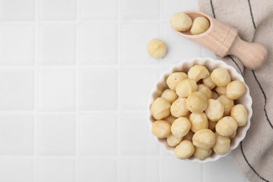 Photo of Bowl and scoop with peeled Macadamia nuts on white tiled table, flat lay. Space for text