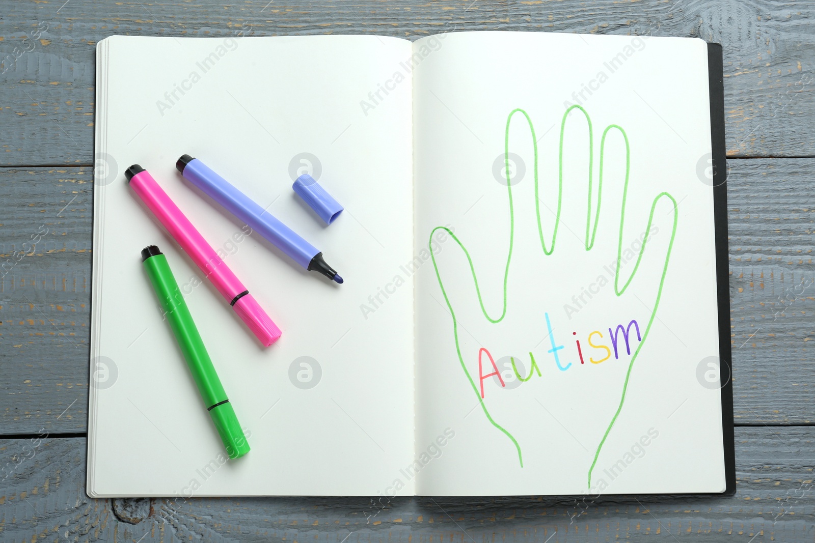 Photo of Colorful felt tip pens and notebook with word Autism on grey wooden table, top view