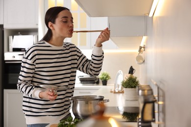 Beautiful woman with wooden spoon tasting soup in kitchen