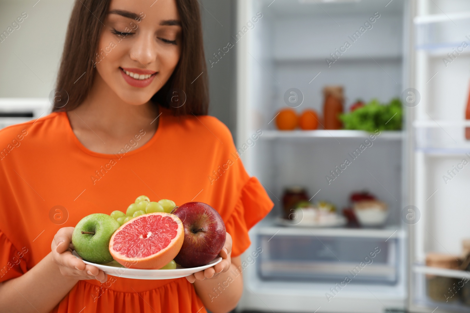 Photo of Concept of choice between healthy and junk food. Woman holding plate with fruits near refrigerator in kitchen