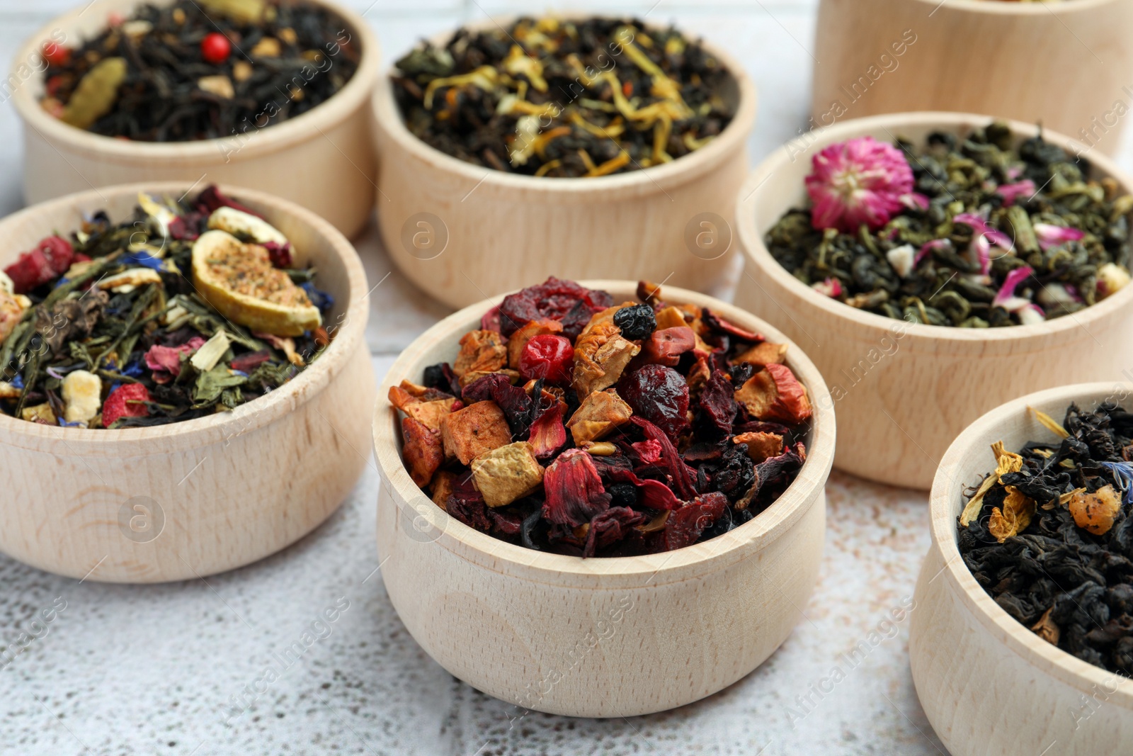 Photo of Different kinds of dry herbal tea in wooden bowls on white tiled surface
