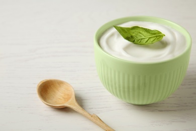 Bowl of fresh sour cream with basil and spoon on white wooden table, space for text