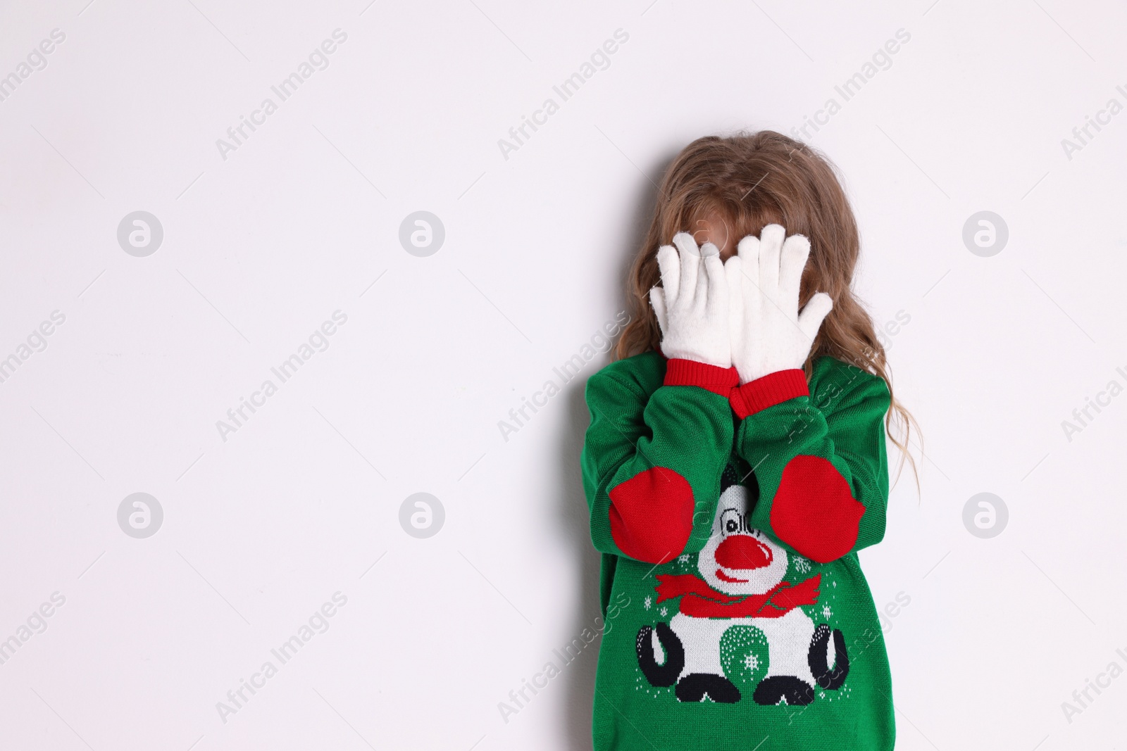 Photo of Cute little girl in green Christmas sweater covering her face against white background