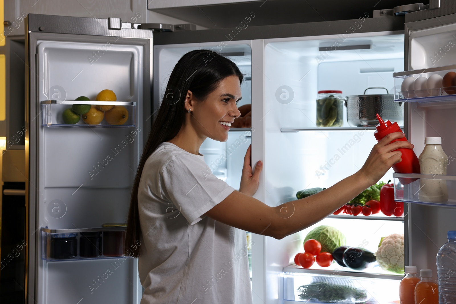 Photo of Young woman taking bottle of ketchup out of refrigerator in kitchen at night