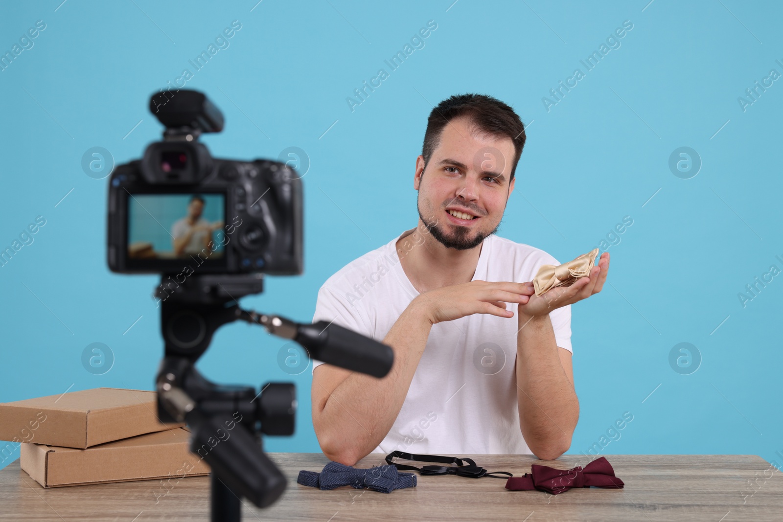 Photo of Smiling fashion blogger showing bow ties while recording video at table against light blue background