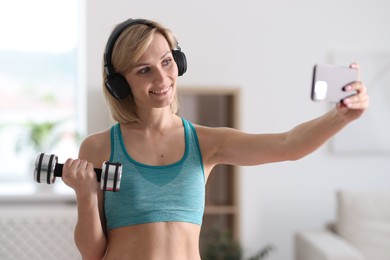 Smiling sports blogger holding dumbbell while streaming online fitness lesson with smartphone at home