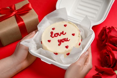 Woman holding takeaway box with bento cake at red table, closeup. St. Valentine's day surprise