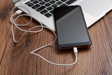 Smartphone connected with charge cable to laptop on wooden table, closeup. Space for text