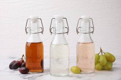 Photo of Different types of vinegar in bottles and grapes on light marble table, closeup