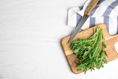 Photo of Fresh arugula, cutting board and knife on white wooden table, flat lay. Space for text