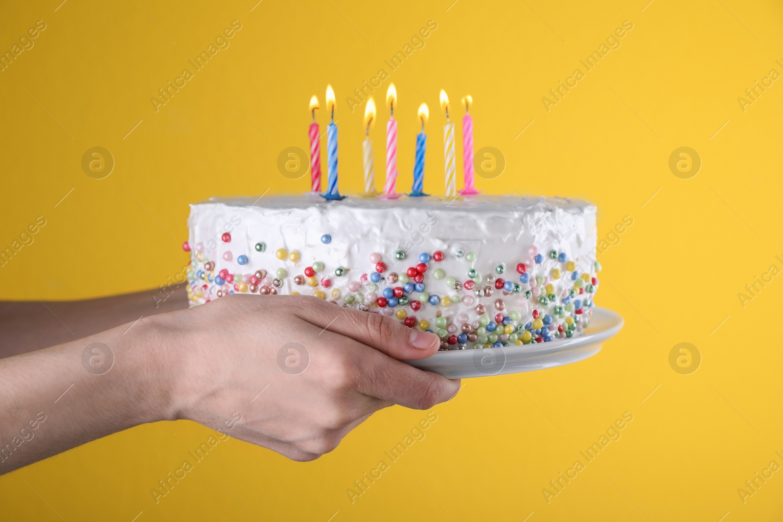 Photo of Woman holding birthday cake with burning candles on yellow background, closeup