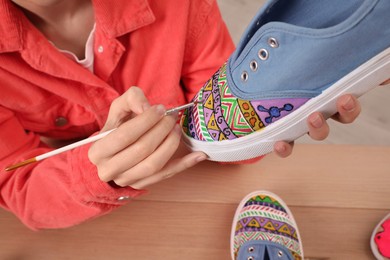 Photo of Woman painting on sneaker at wooden table, closeup. Customized shoes