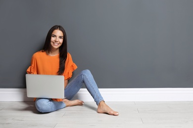 Young woman with modern laptop sitting on floor near grey wall