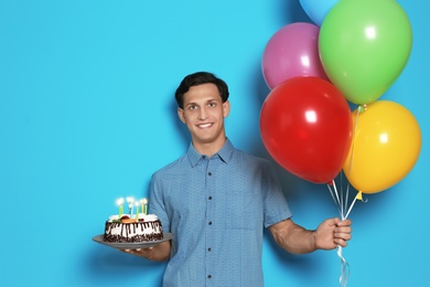 Young man with birthday cake and bright balloons on color background