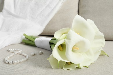 Photo of Beautiful calla lily flowers tied with ribbon, wedding dress and jewelry on sofa